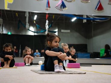Young girls sitting on mats in a gym, practicing a seated exercise.