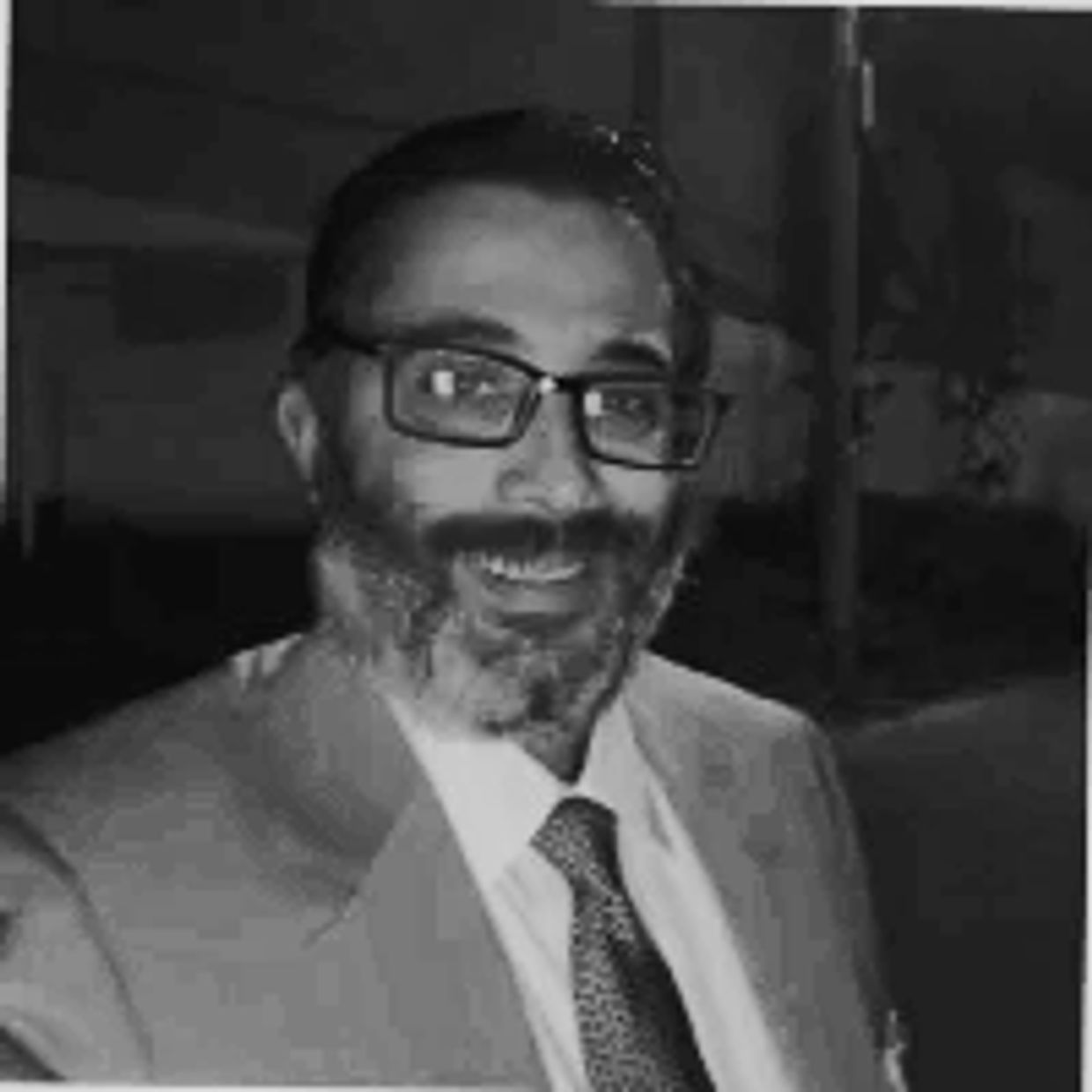 Smiling man with glasses, beard, and suit in a black-and-white photo.
