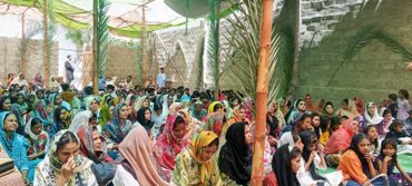 Large group of women and children seated under a canopy during a community gathering.