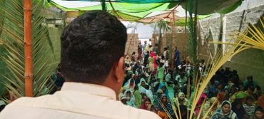 Man addressing a large seated crowd under a makeshift canopy.