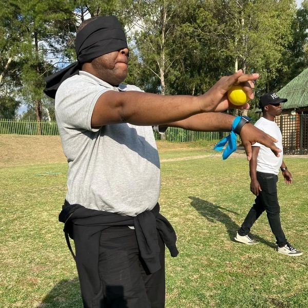 Blindfolded man holding a yellow ball with another person walking nearby outdoors.