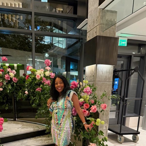 Woman in a shimmering dress posing happily beside vibrant pink flowers indoors.