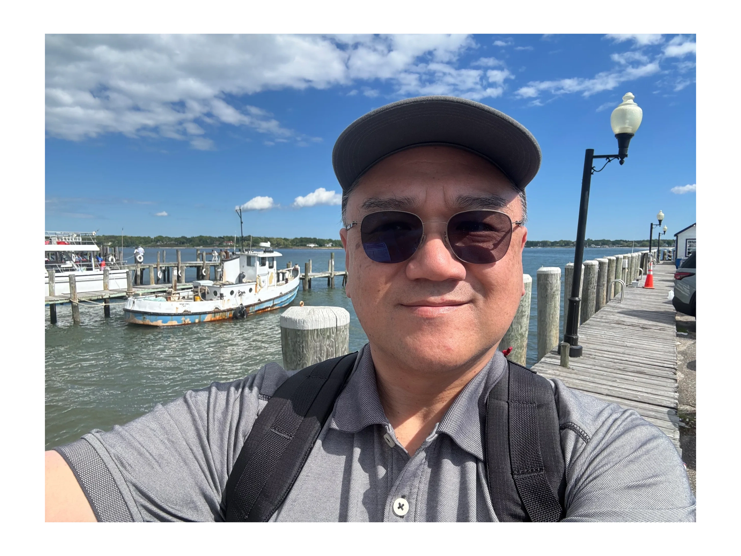 Man wearing sunglasses and a cap takes a selfie on a sunny dock with boats in the background.