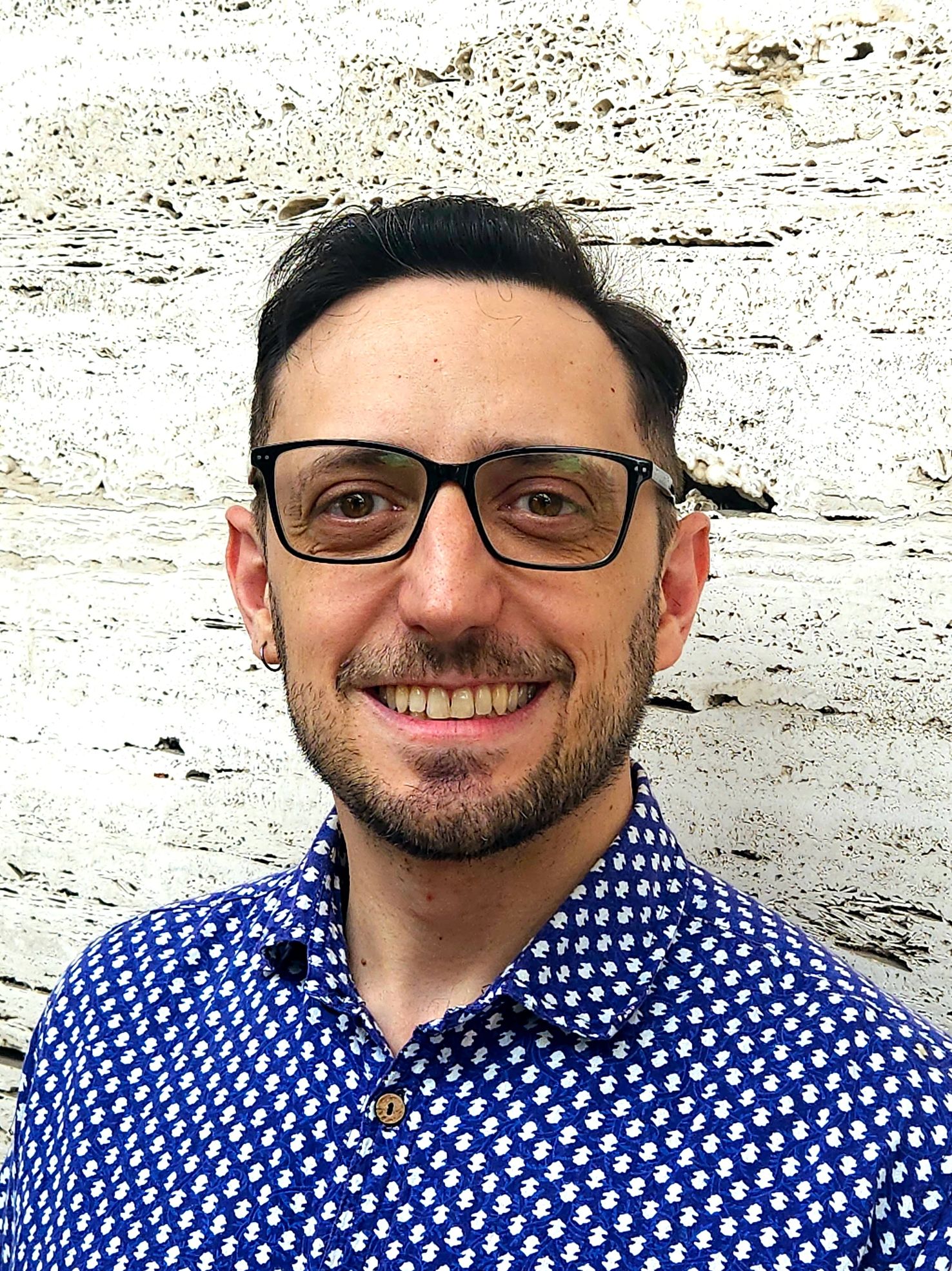 Smiling man with glasses wearing a blue patterned shirt against a textured wall.