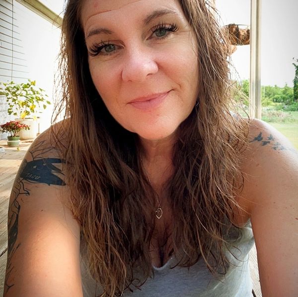 Woman with long hair and tattoos smiling indoors with plants in background.