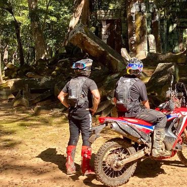 Two motorcyclists in gear at an ancient stone ruin in a forest.