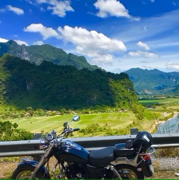 Two motorcycles parked with a scenic mountain and river backdrop under a blue sky.
