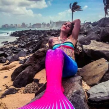 A woman in a pink and blue mermaid tail reclined back over a rocky shoreline.
