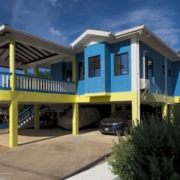 A vibrant blue and yellow raised house with parked cars underneath.