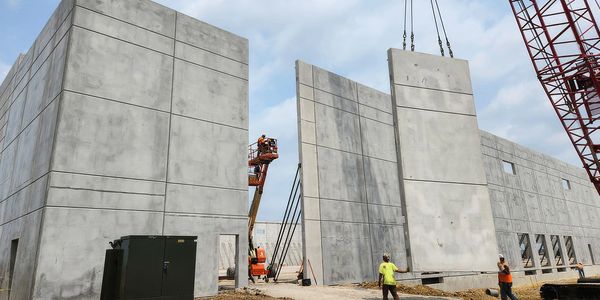Workers guide large concrete panels being lifted by a crane on a construction site.