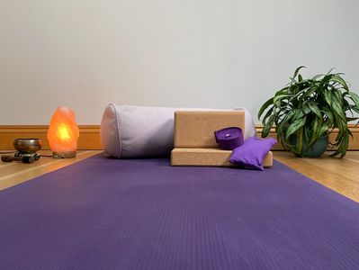 A purple yoga mat rolled out with yoga props on top, next to an orange lamp and green house plant.