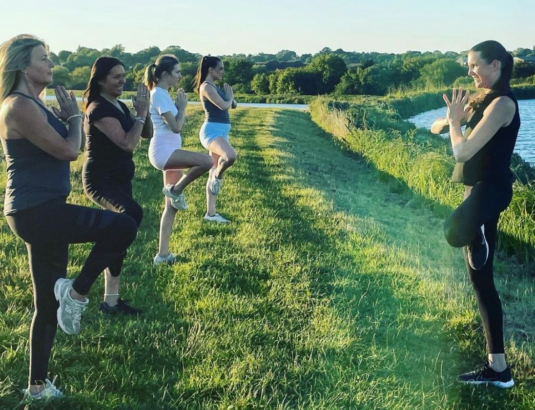 5 woman practicing yoga in a field, standing in tree pose and smiling.