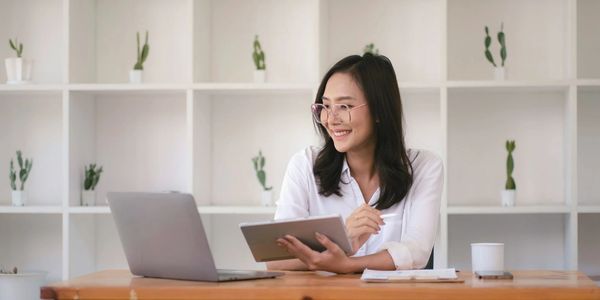 A woman sitting at a desk smiling while holding a tablet and looking at her laptop.
