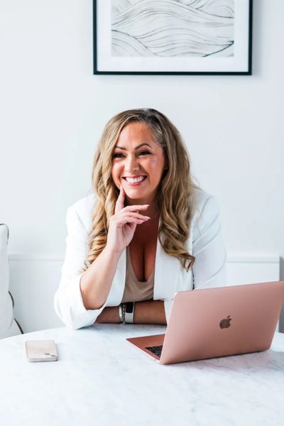Smiling woman with blonde hair working on a rose gold laptop at a white table.