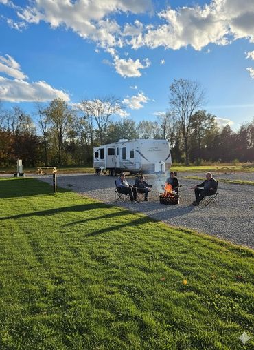 Four people sitting around a campfire near a camper in a sunny outdoor setting.