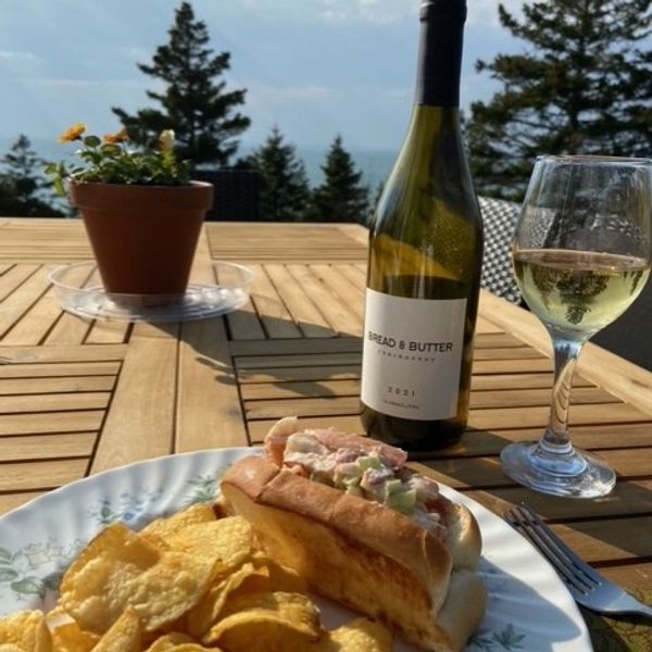 Outdoor meal with lobster roll, chips, and white wine on a wooden table.