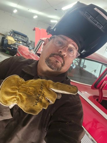 Rodney wearing welding helmet and glove in a garage with classic cars.