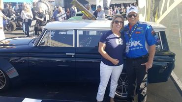 A smiling couple poses beside a classic blue American car at a crowded outdoor event.