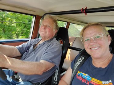 Two smiling people inside a car wearing racing harnesses.