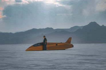 A man stands beside a sleek, orange speed vehicle on a vast salt flat under a cloudy sky.