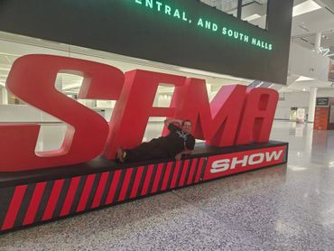 Man lounging on large red "SEMA SHOW" letters inside a spacious convention center.