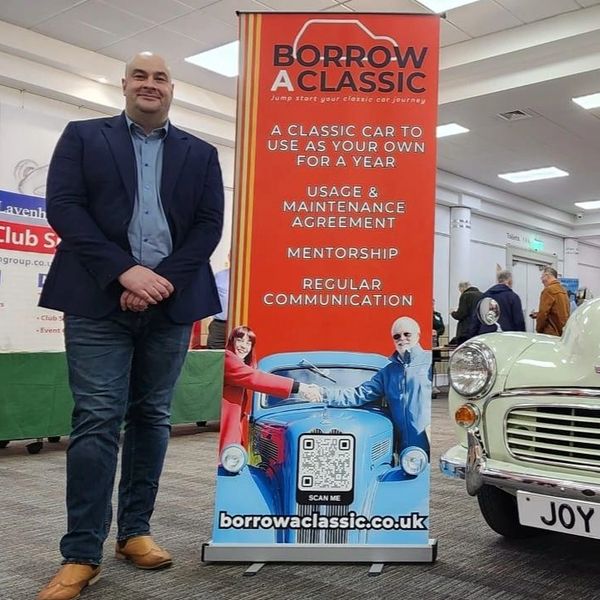 Man standing next to a banner promoting classic car borrowing for a year.