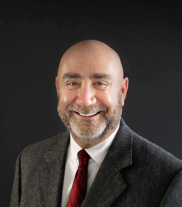 Smiling bald man with beard in a suit and red tie against a dark background.