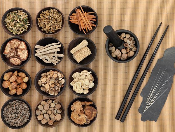 Bowls of herbs sit on a bamboo mat next to chopsticks and acupuncture needles.