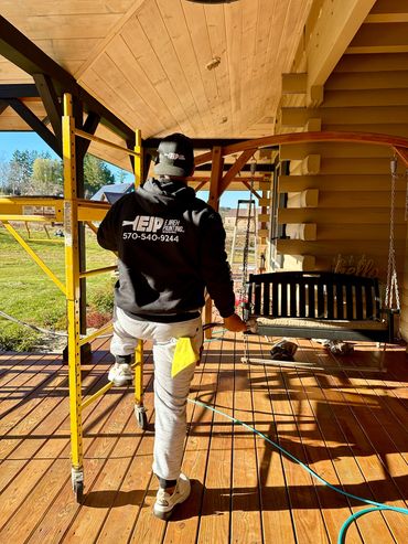 A painter steps onto scaffolding on a sunny porch, wearing branded workwear.