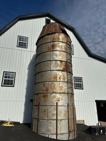 Rusty metal silo in front of a white barn under a cloudy sky.