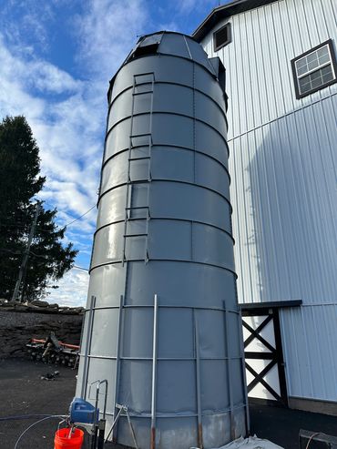 A tall, gray metal silo with a ladder attached, next to a white barn under a partly cloudy sky.