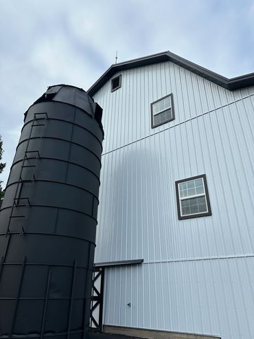 Black silo next to a large white barn with windows under a cloudy sky.