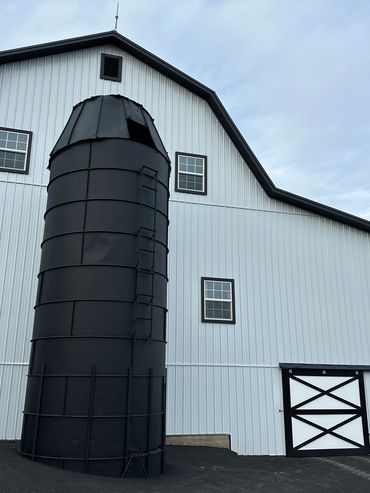 Black silo stands beside a white barn with black trim on a cloudy day.