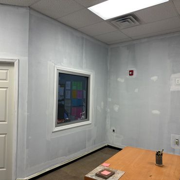 Office room with patchy gray walls, wooden tables, and a window to another office.