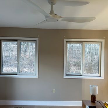 Room with three windows, beige walls, ceiling fan, and a wooden side table with a lamp.
