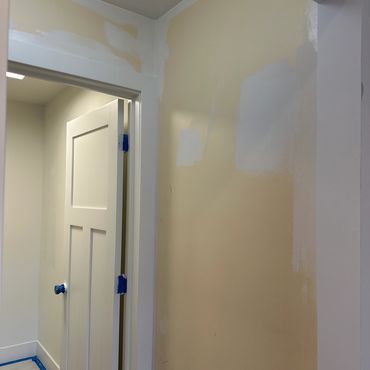 Wall partially painted with white patches over beige, door with blue tape on hinges and knob.