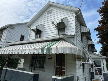 White two-story house with green-striped metal awnings and a ladder beside it.