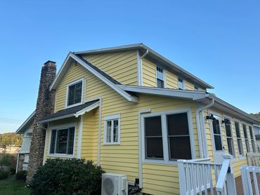 Yellow house with white trim and stone chimney under a clear blue sky.
