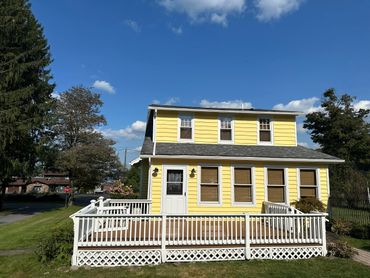 Bright yellow house with white trim and a spacious front porch under a clear blue sky.