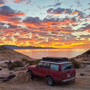 A red off-road vehicle parked at sunset by a calm body of water and hills.