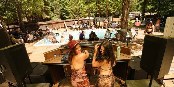 Two women DJing at a lively outdoor pool party in a sunny backyard.
