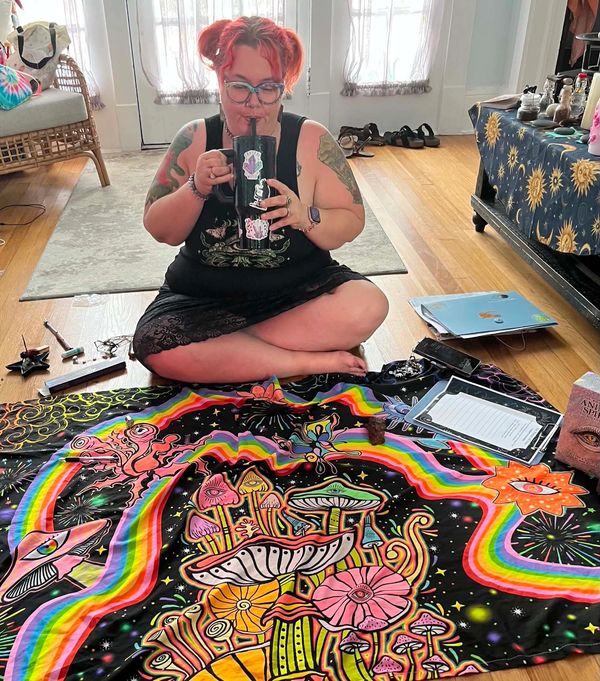 Person with red hair sitting cross-legged, drinking from a decorated mug over a colorful psychedelic tapestry.