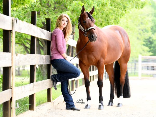 Woman in a pink sweater leaning on a fence next to a brown horse.