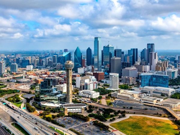 A vibrant cityscape of Dallas with skyscrapers and highways under a partly cloudy sky.