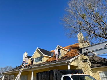 Workers removing old shingles from a yellow house roof under a clear blue sky.