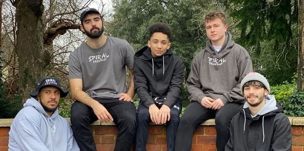 Five young men posing casually outdoors in hoodies and caps.