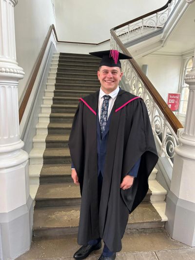 Young man in graduation gown standing on staircase, smiling.