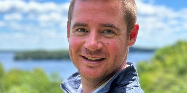 Smiling man outdoors with greenery and water in background.
