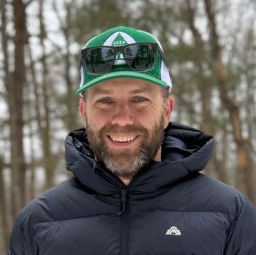 Smiling man in a green cap and black jacket standing outdoors in a forest.