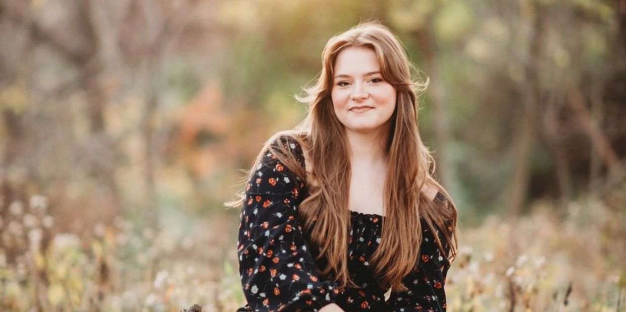 Young woman with long hair smiles outdoors in a floral dress.